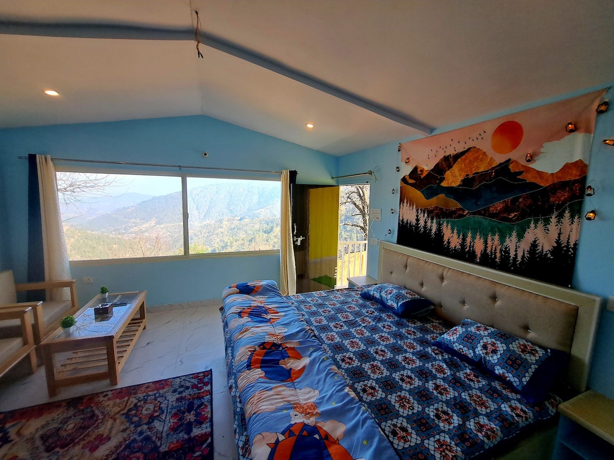 A spacious room features a large window presenting sweeping views of the mountains and valley. The bed, adorned with a colorful patterned bedspread, is complemented by natural light filtering through. A cozy sitting area includes two chairs and a coffee table, contributing to the inviting ambiance.