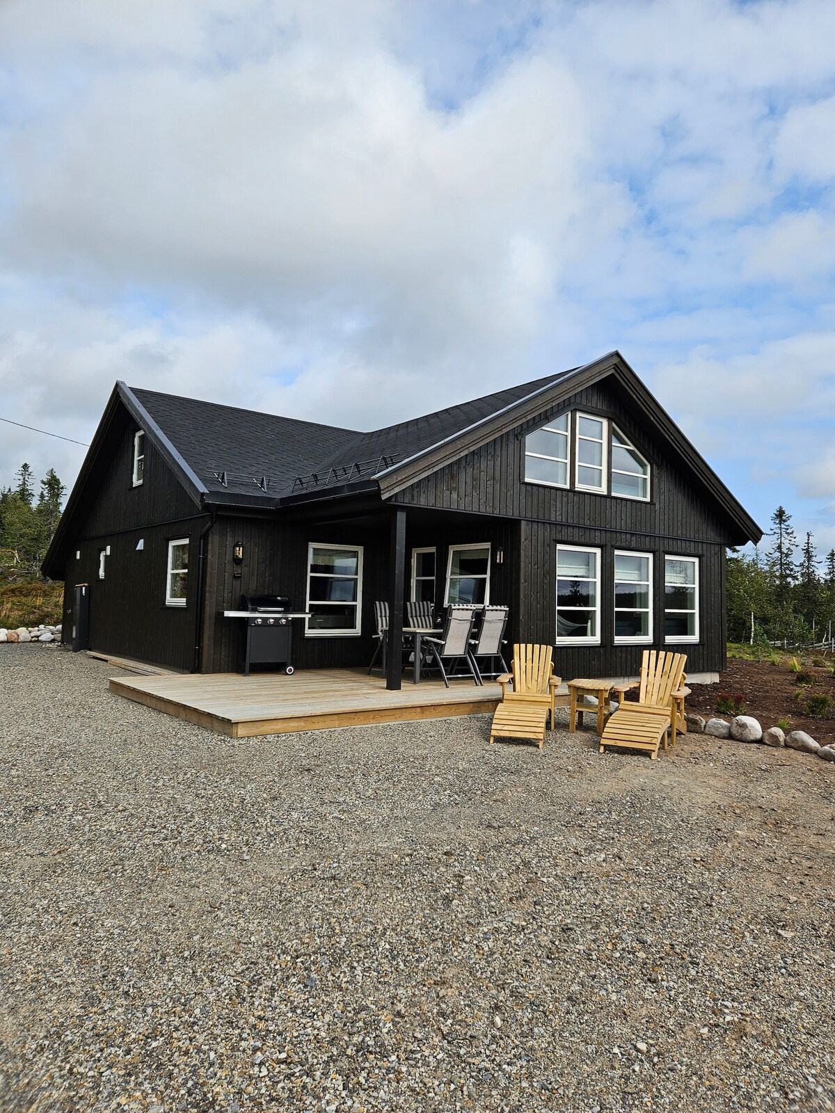 The two-story cabin features a dark wooden exterior and large windows that provide ample natural light. A spacious deck is equipped with a dining set and two wooden lounge chairs. The surrounding area includes gravel pathways and greenery.