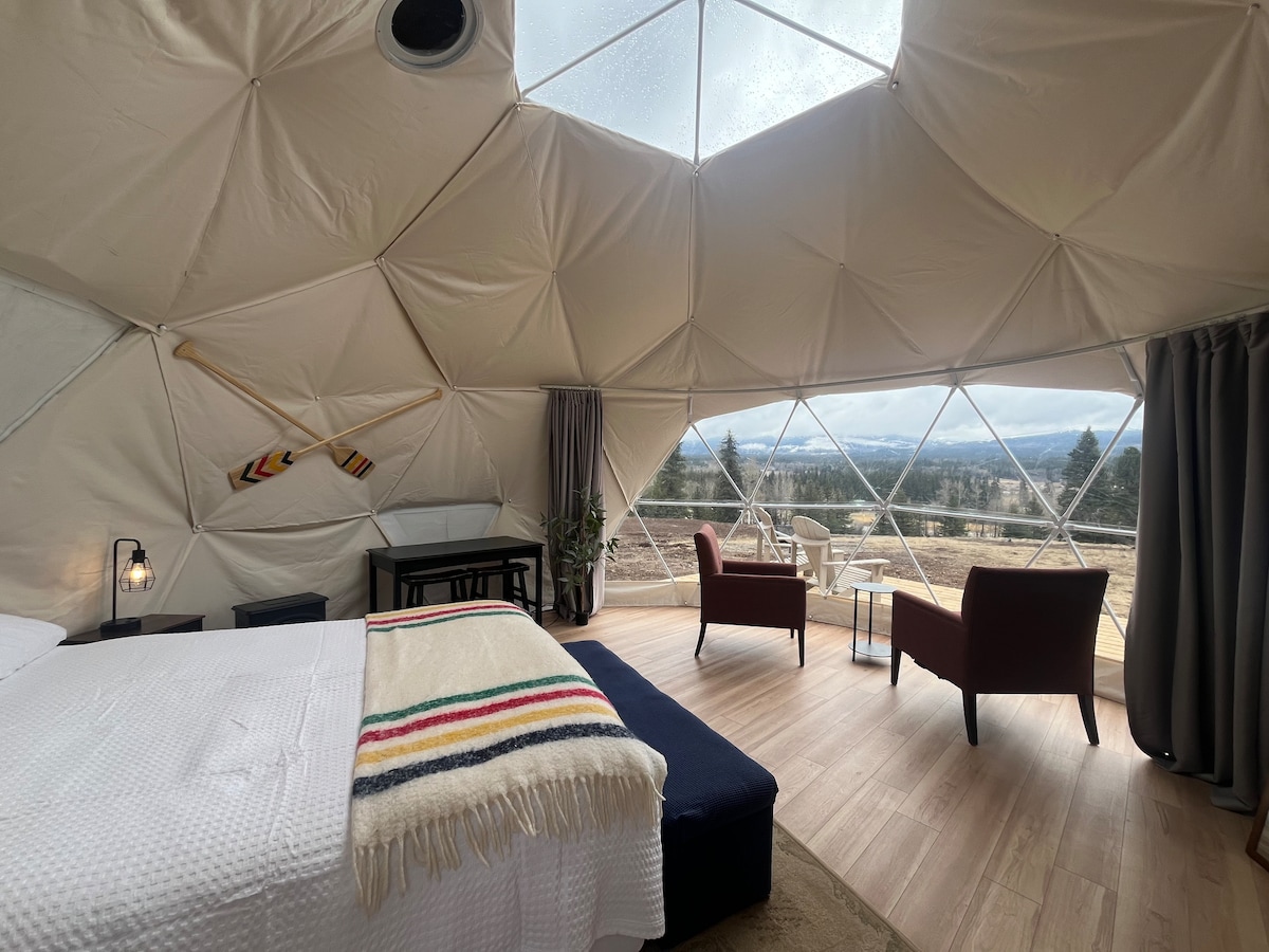 The interior of the glamping dome features a queen bed covered with a striped blanket. A seating area with two chairs and a small table is positioned near large windows, offering expansive views of the landscape. Natural light filters through the dome, enhancing the spacious ambiance.