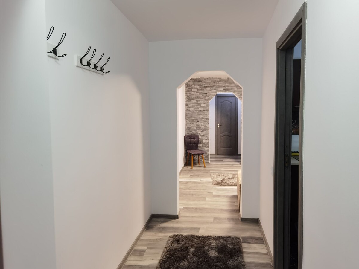 A well-lit hallway features a smooth wooden floor and white walls, accented by a textured stone wall in the background. Wall hooks are mounted on the left for convenience, and a small chair is positioned in the distance, inviting guests to relax.