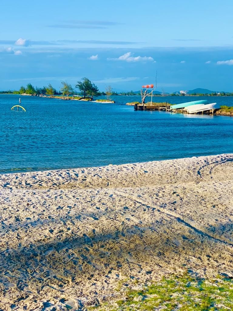 A serene view of a calm lagoon features gentle waves lapping against a sandy shoreline. A small boat is anchored nearby, and distant islands are visible against a bright blue sky, completing the tranquil coastal landscape.