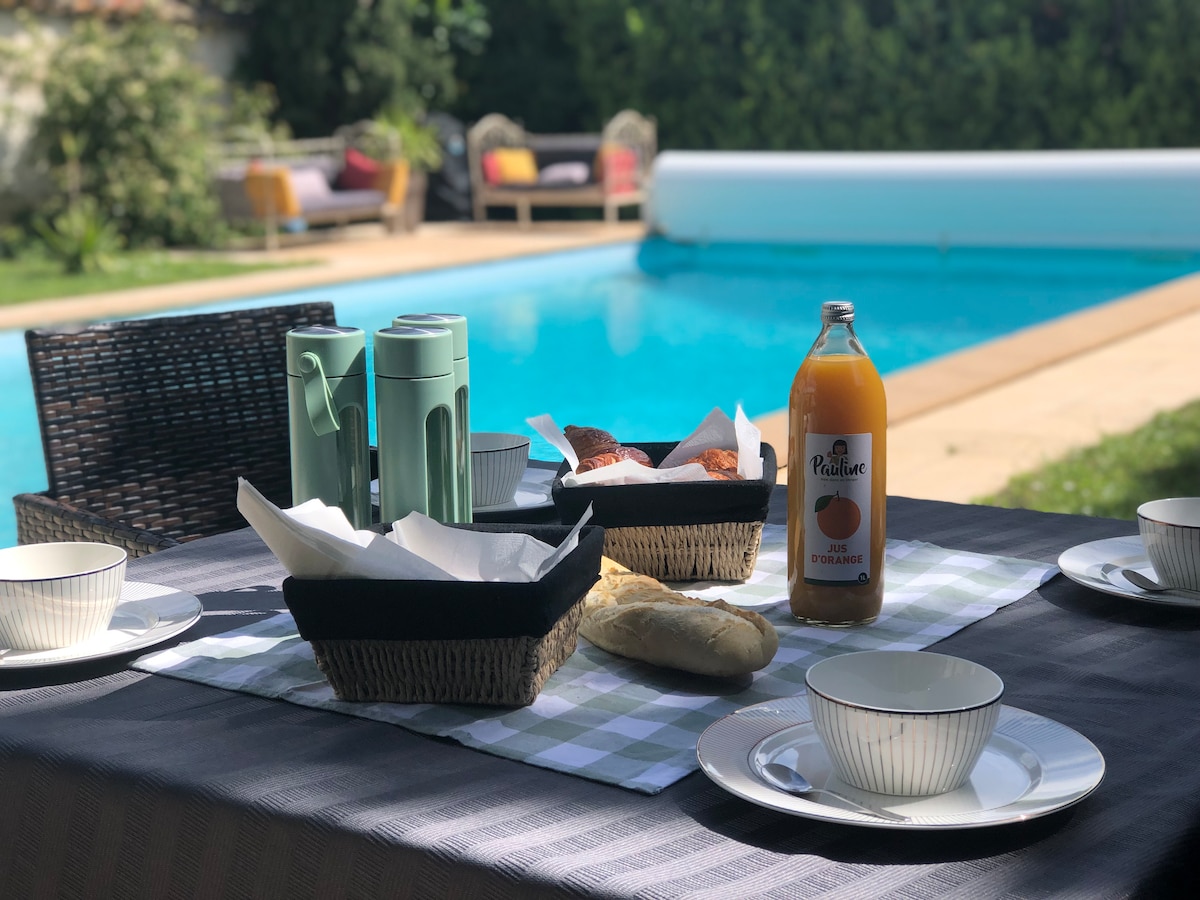 A dining table is set outdoors, featuring four bowls, plates, and napkins. Two baskets contain baked goods, alongside a bottle of orange juice. In the background, a swimming pool reflects natural light, complemented by green foliage and comfortable seating areas nearby.