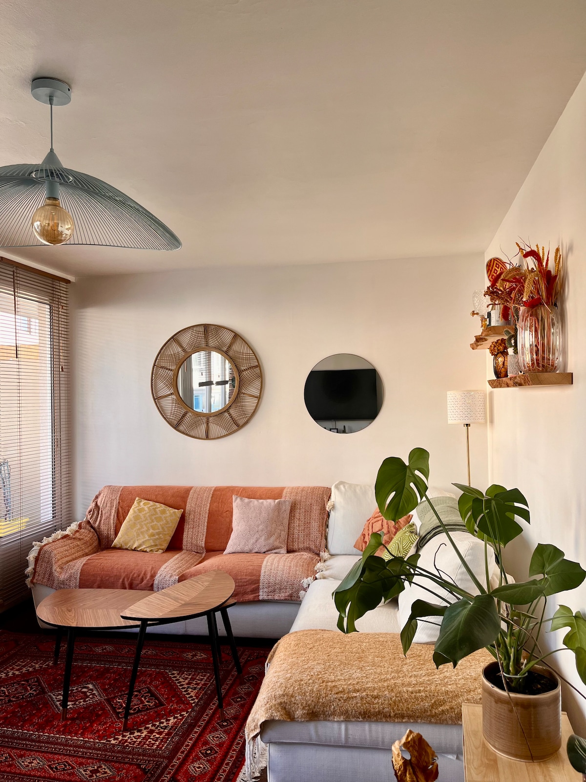 A cozy living area is furnished with a light-colored sofa adorned with throw pillows. A pair of decorative mirrors reflect natural light, while a small table sits between seating. A potted plant adds a touch of greenery, complemented by a warm rug beneath.