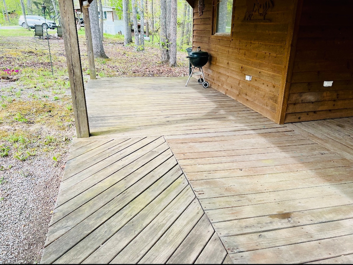 A spacious wooden deck area is viewed, featuring a grill placed on one side. The natural wood grain of the flooring is visible, surrounded by a wooded landscape. Sunlight filters through the trees, casting gentle shadows across the deck.
