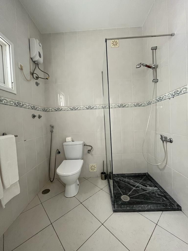 A clean and modern bathroom features a walk-in shower enclosed by glass, with sleek fixtures mounted on the wall. A white toilet is positioned near the corner. The tile floor is complemented by light-colored walls, and the shower area showcases dark marble accents.