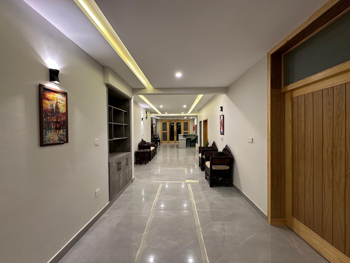 A wide corridor is presented, featuring shiny marble flooring and neutral-colored walls. Artwork is hung on the walls, and seating arrangements with dark wood furniture are positioned along the sides. Warm lighting is provided by wall-mounted fixtures, enhancing the spacious feel.