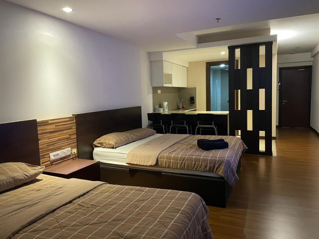 A spacious guest room is highlighted by two neatly made beds with neutral-colored bedding. A kitchenette area can be seen in the background, featuring modern appliances and seating for two. Soft lighting enhances the warm tones of the wooden flooring.