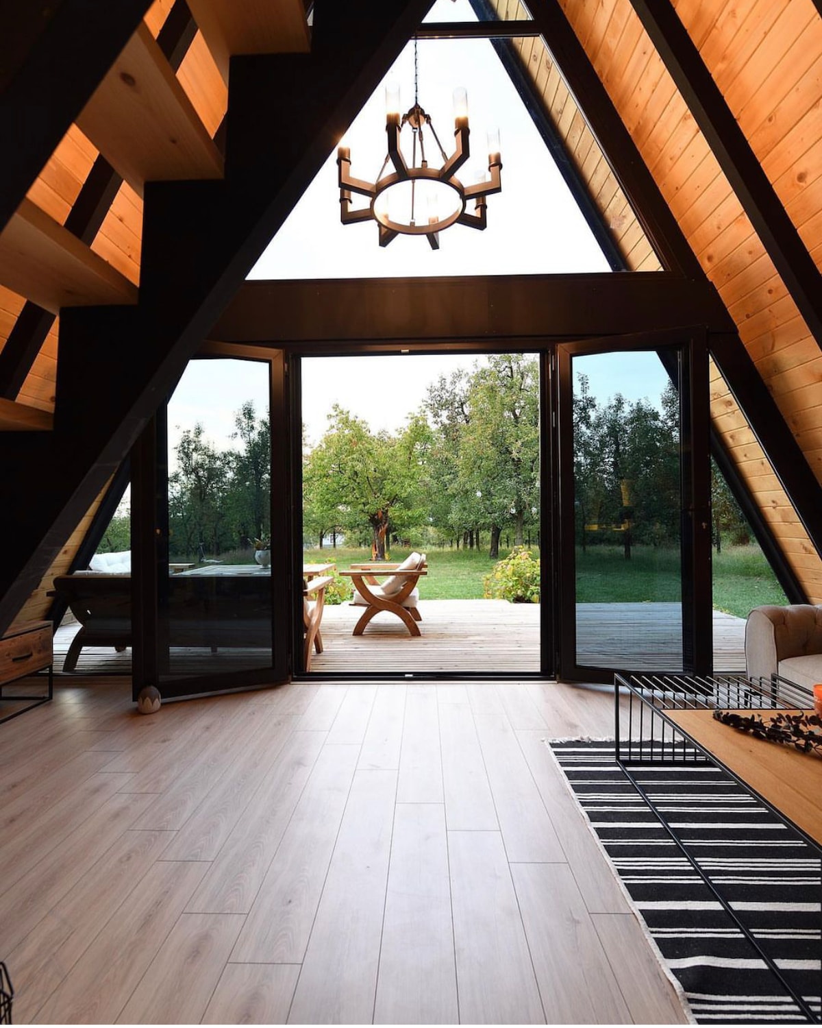 A modern A-frame structure features large glass doors that open to a grassy area beyond. Natural wood beams and paneling are highlighted within the space. A light fixture hangs from the ceiling, and a striped rug is placed on the wooden floor.