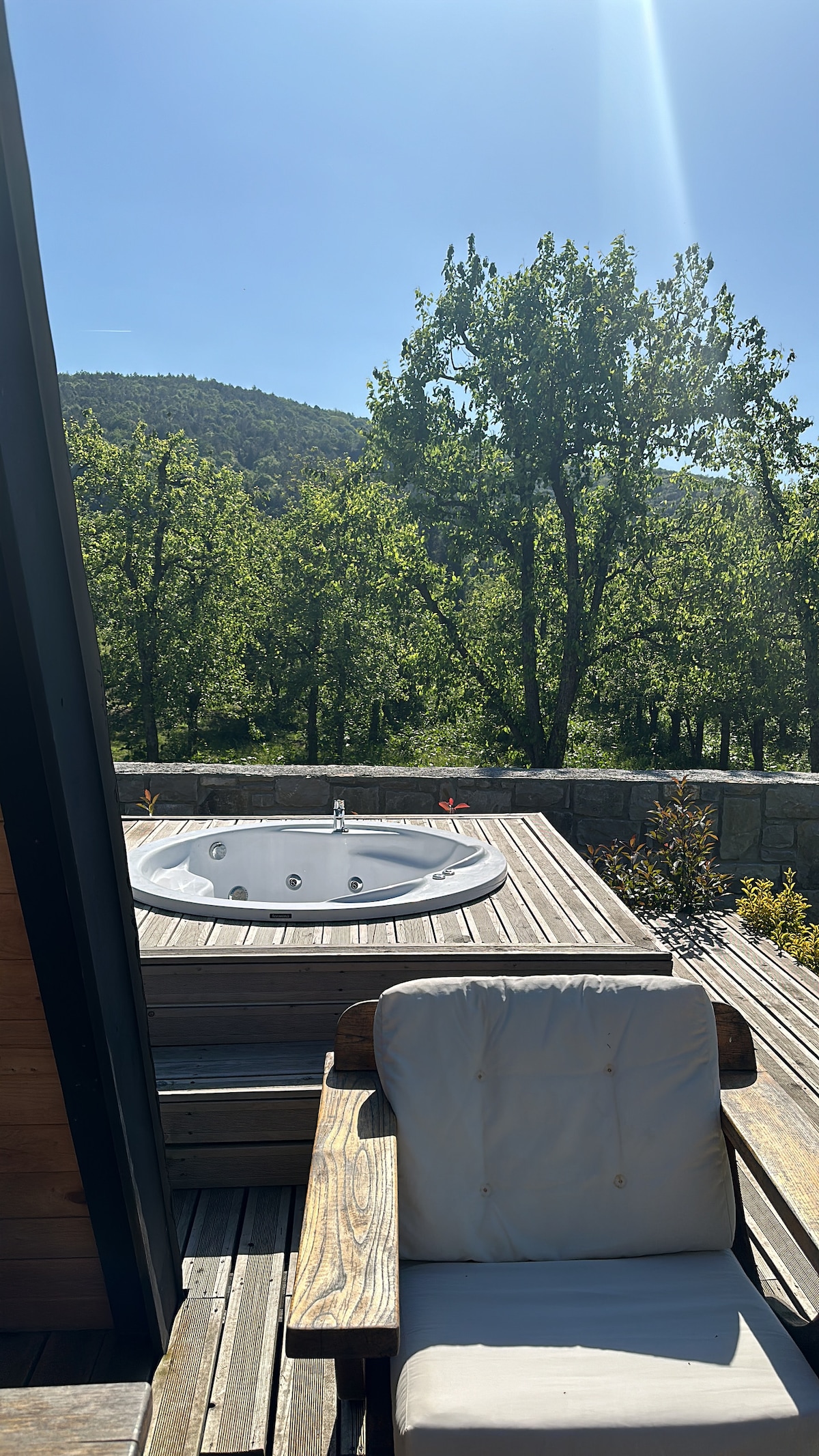 A hot tub is positioned on a wooden deck, surrounded by lush greenery and towering trees. Sunlight casts a warm glow over the space, enhancing the natural beauty of the outdoor area, which offers a serene view of the surrounding landscape.