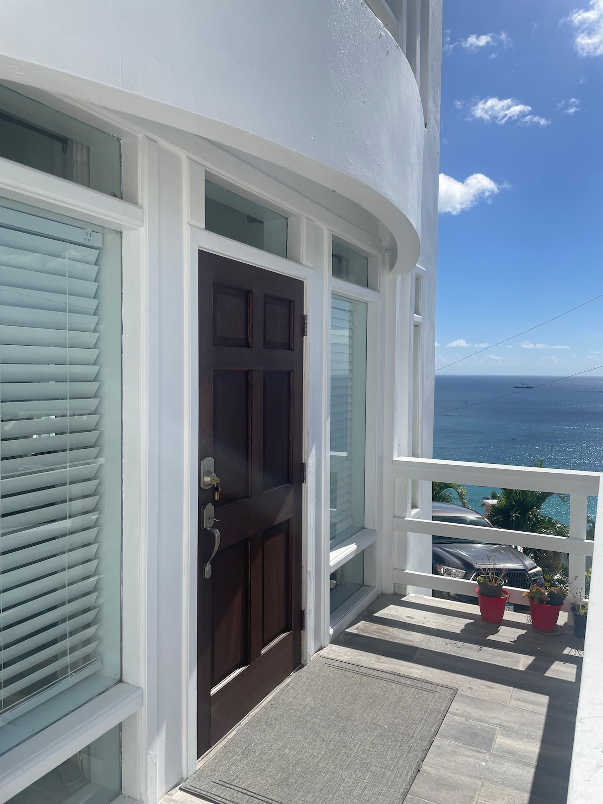 An entrance to the unit features a wooden door framed by large windows. Light filtering through the blinds creates an airy feel. A small rug lies at the threshold, while colorful potted plants add a touch of greenery. The ocean is visible in the background under a clear sky.