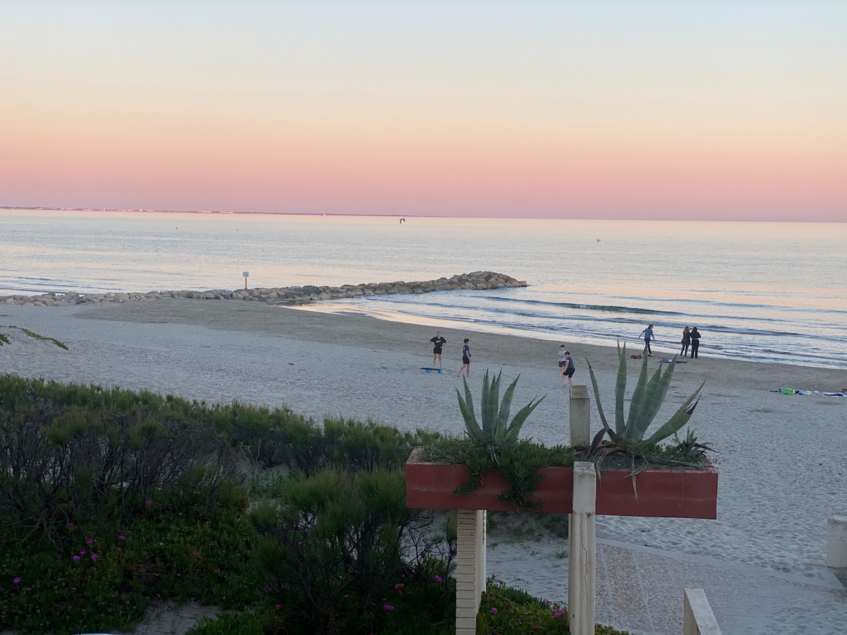 A serene beachfront view captures soft, pastel colors in the sky at sunset. Gentle waves lap against the shore, where small groups can be seen enjoying the beach. Lush greenery frames the foreground, adding natural beauty to the coastal scene.