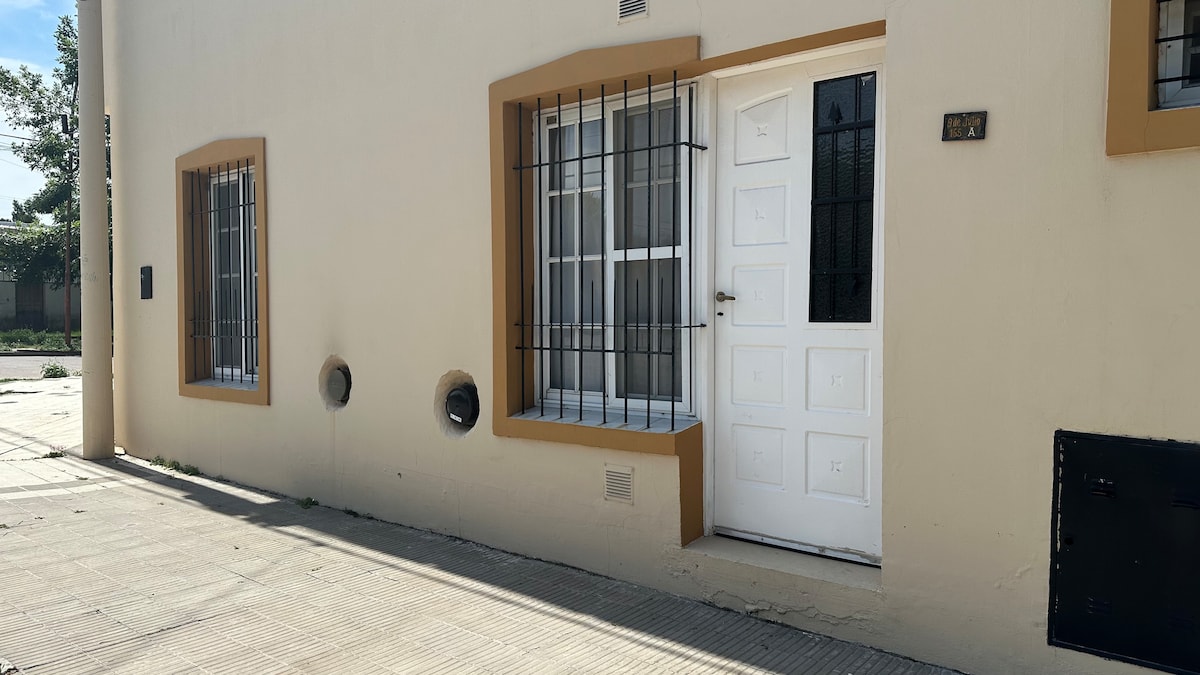 The exterior of a modest building is visible, featuring a white door with a simple design and window bars for security. Two small round ventilation openings are present on the wall, alongside neatly framed windows that allow natural light.