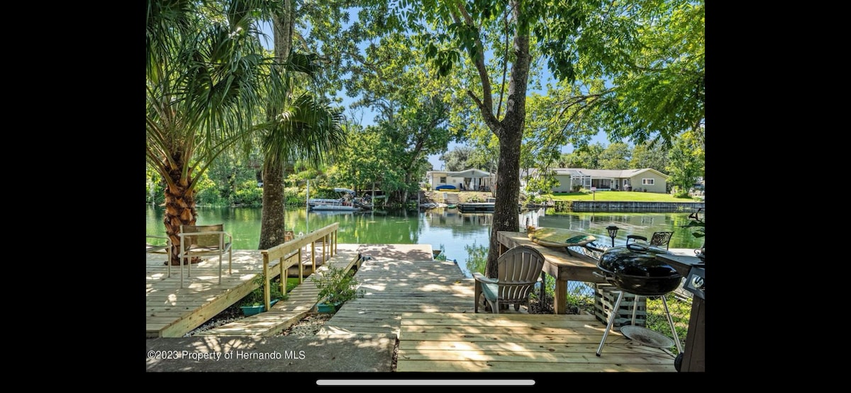 A wooden deck borders a peaceful freshwater canal, surrounded by lush trees. A pair of chairs and a table are positioned to the side, with a grill nearby. The calm water reflects the greenery, creating a tranquil atmosphere for relaxation.