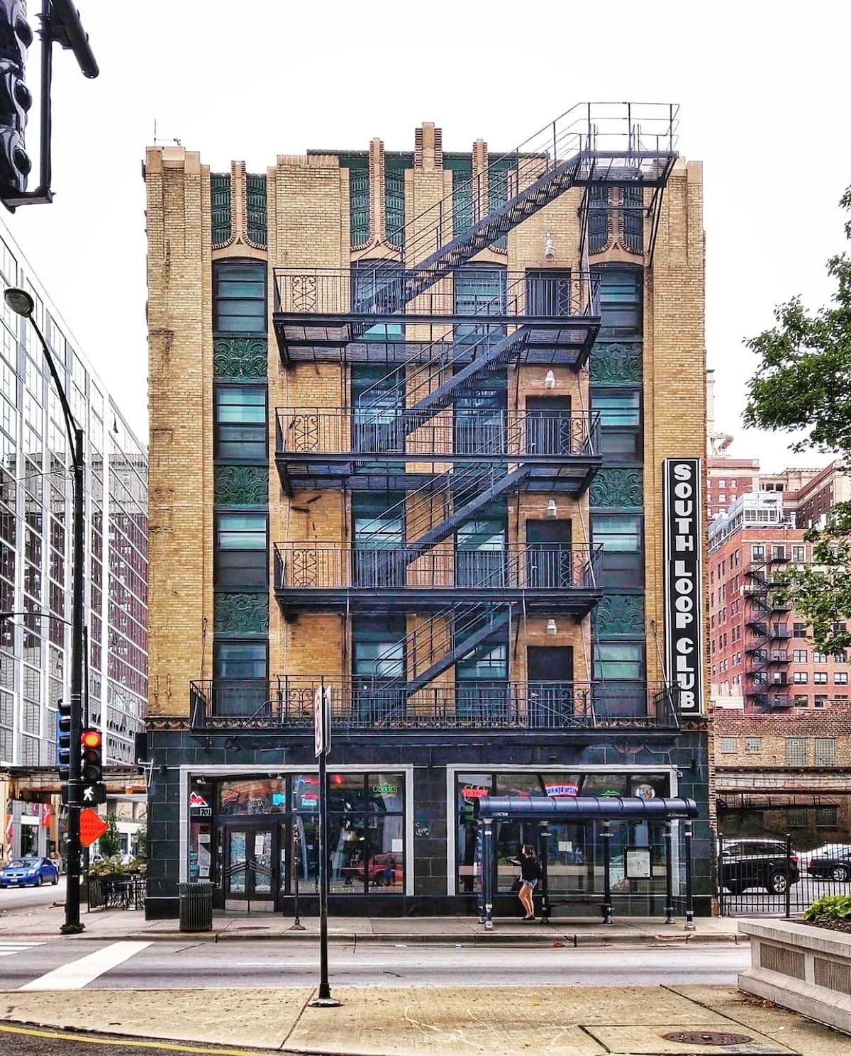 A five-story building characterized by its vintage brick facade and decorative ironwork. An outdoor fire escape extends along the side, while large glass windows offer views of the bustling street below. A prominent sign displays the name 'South Loop Club.'