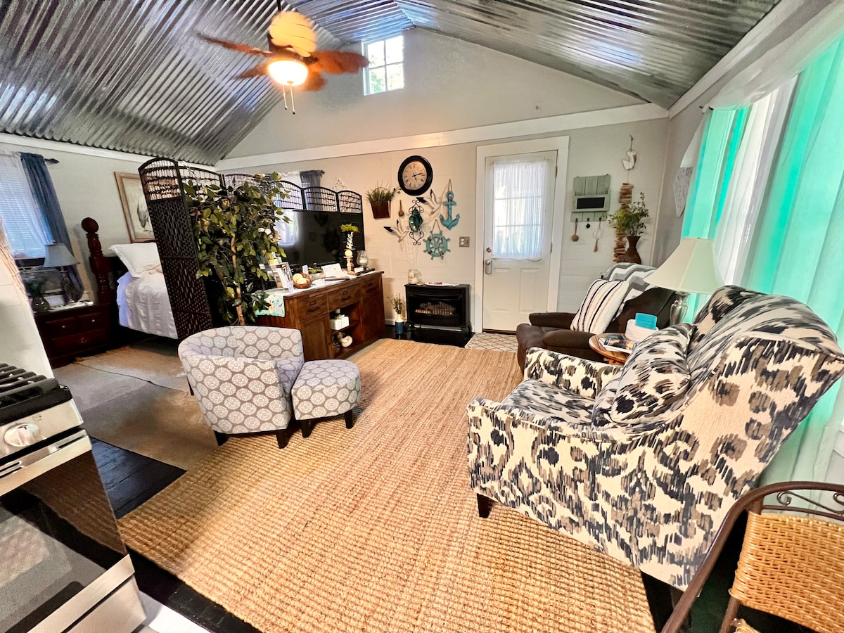 An open living space features a cozy seating area with patterned chairs and a small table. A decorative screen partially divides the room, with a simple entertainment unit and a fireplace nearby. The ceiling is lined with tin panels, and colorful curtains frame the windows.
