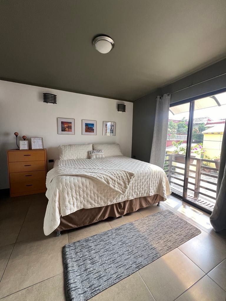 A bedroom is furnished with a spacious bed, complemented by light-colored bedding and decorative pillows. A chest of drawers is positioned nearby, while framed artwork adorns the walls. Natural light enters through a sliding door, which leads to a small balcony area.