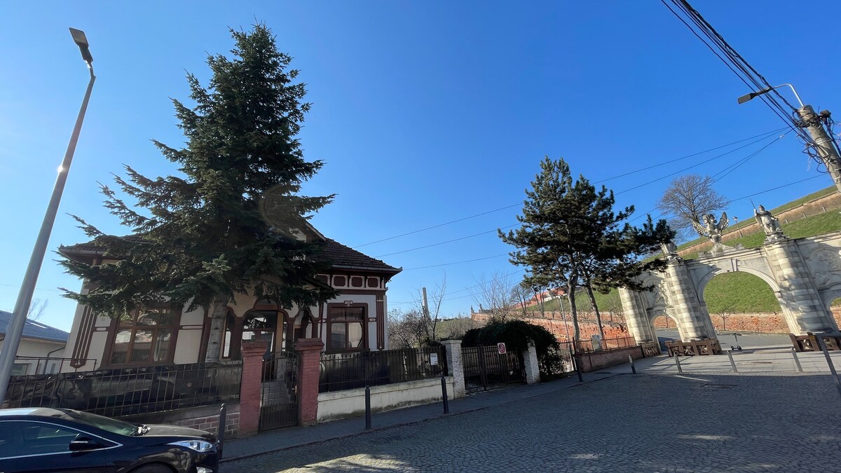A historic building is featured with a traditional architectural style, set against a clear blue sky. Lush green trees surround the structure, while a cobblestone street leads to the entrance. Nearby fortifications and a hillside are visible, adding to the location's unique charm.