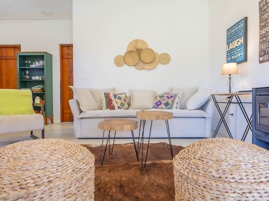 A comfortable living area features a neutral sofa adorned with decorative pillows. Two round wooden tables serve as a centerpiece on a textured area rug. Natural light brightens the space, accented by warm wood tones from the surrounding furniture.