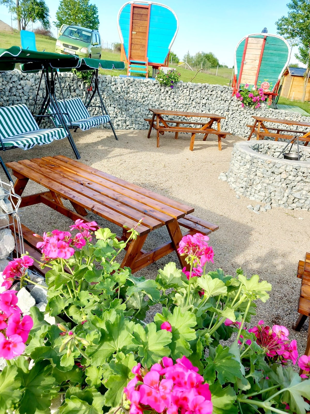 A relaxing outdoor area features wooden picnic tables surrounded by vibrant pink flowers. Comfortable lounge chairs with striped cushions are positioned under a canopy. Stone walls define the space, and brightly colored structures add a playful touch to the landscape.