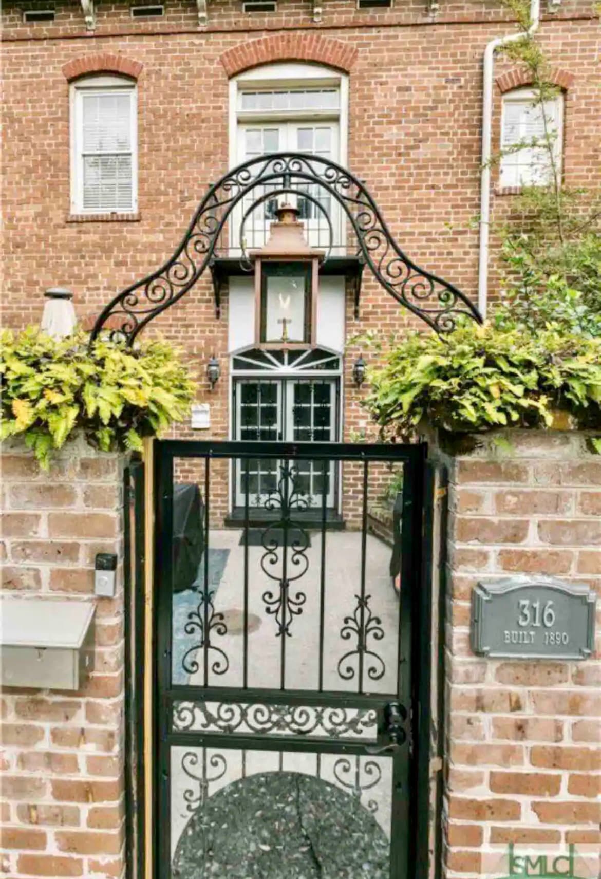 An ornate wrought-iron gate leads into a charming courtyard, framed by brick walls adorned with greenery. The gate features delicate scrolling designs, while above it hangs a gas lantern. The townhouse number '316' is prominently displayed beside the gate.