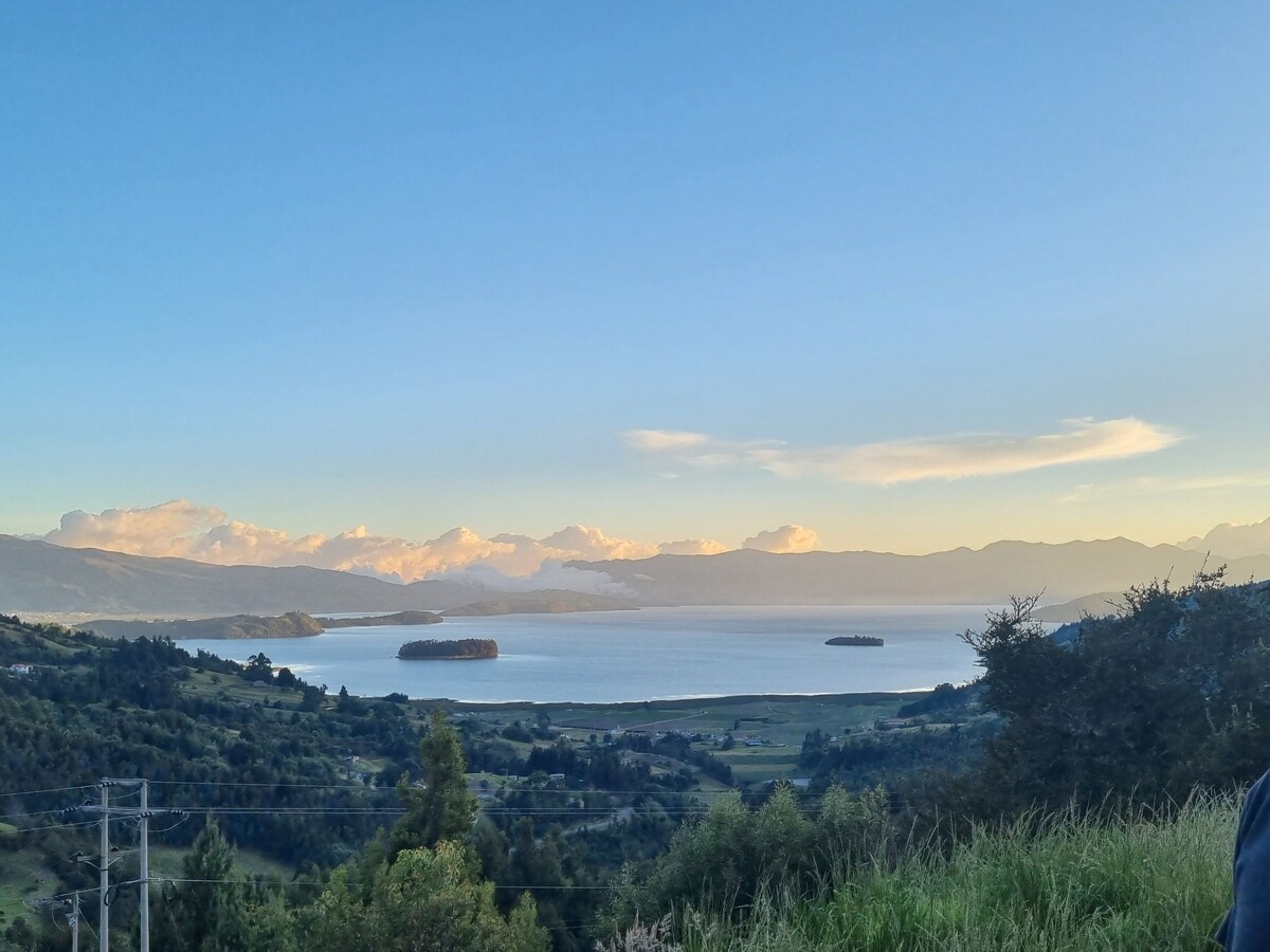 A panoramic view of Laguna de Tota is captured, showcasing calm waters reflecting the sky. Surrounded by lush greenery, mountains rise in the background, with soft clouds drifting above. The scene offers a serene atmosphere, inviting contemplation of the natural beauty.