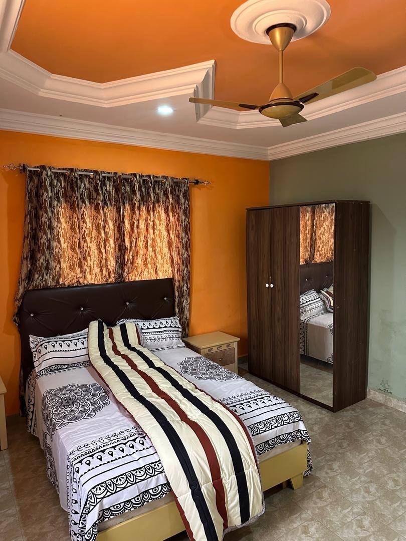 A room is presented with an orange accent wall and contrasting green wall. A spacious bed with decorative bedding is positioned beneath a window adorned with patterned curtains. A reflective wardrobe stands beside the bed, enhancing the room's functionality.