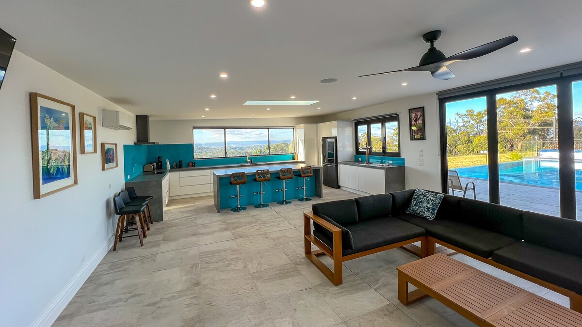 A spacious living area features large windows offering views of the Yarra Valley. Modern furnishings include a sectional sofa and a wooden coffee table. A well-equipped kitchen with a large island and stools sits in the background, complemented by natural light streaming through skylights.