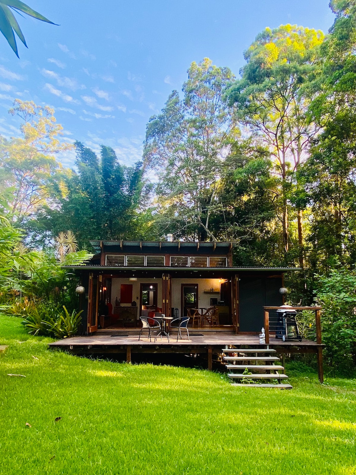 A charming retreat is nestled among lush greenery, featuring a wooden deck with outdoor seating. The structure showcases large glass doors that invite natural light, surrounded by tall trees and vibrant plants, offering a serene connection to nature.
