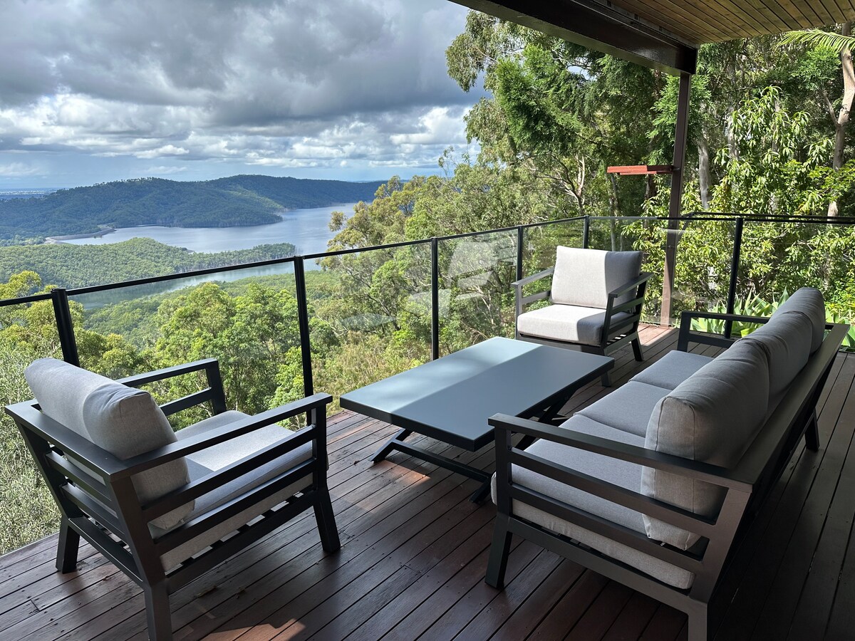 An outdoor seating area features comfortable gray sofas and a matching coffee table, positioned on a wooden deck. The expansive view reveals a forested landscape and a body of water, framed by lush greenery and a partly cloudy sky.