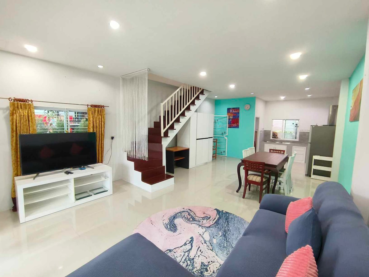 An open living and dining area features a comfortable couch, a television, and a decorative area rug. A staircase leads to an upper level. The dining table and kitchen are visible in the background, complemented by large windows that allow natural light to fill the space.