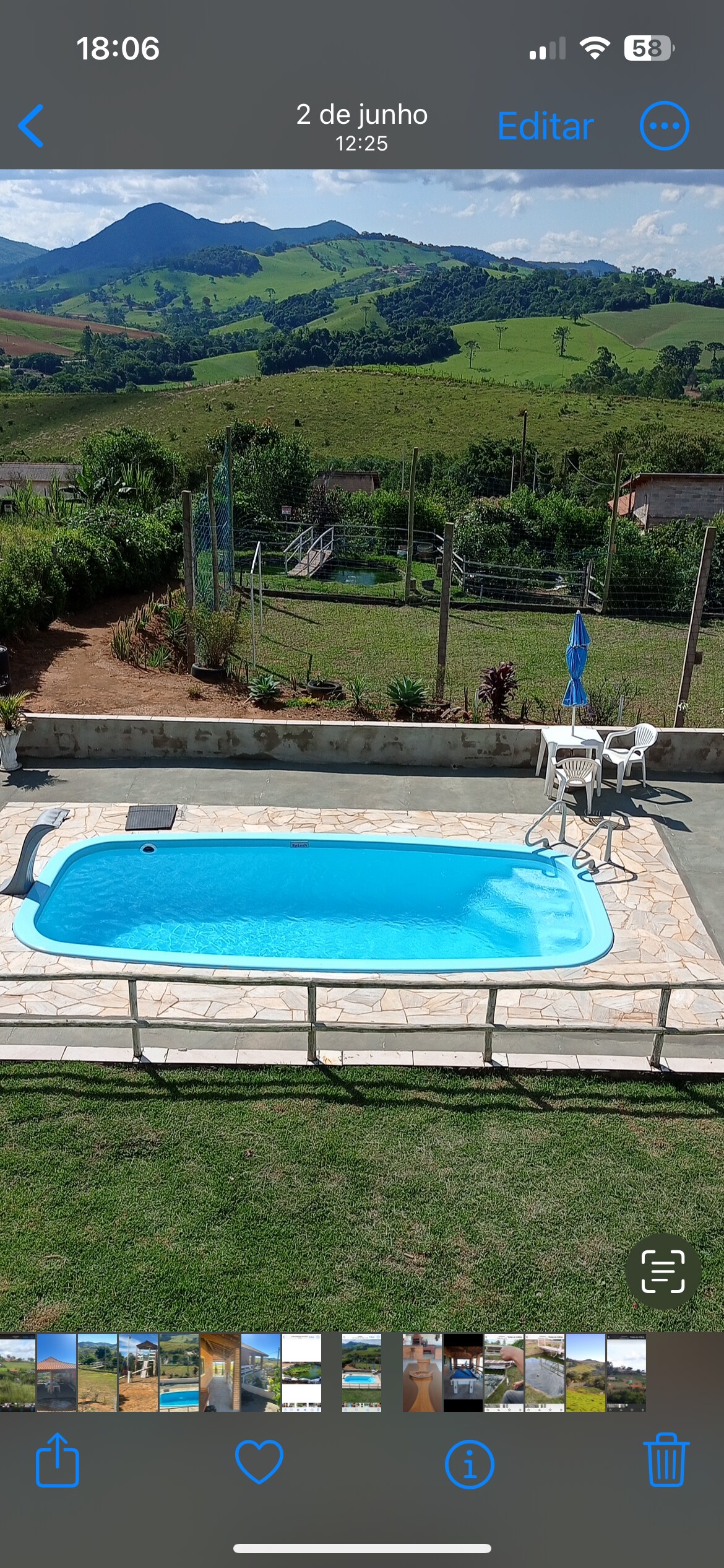An outdoor swimming pool is surrounded by a paved patio, with a selection of lounge chairs positioned nearby. Lush green hills and distant mountains frame the landscape, offering a serene backdrop for relaxation. A vibrant blue umbrella stands next to the pool, providing shade.