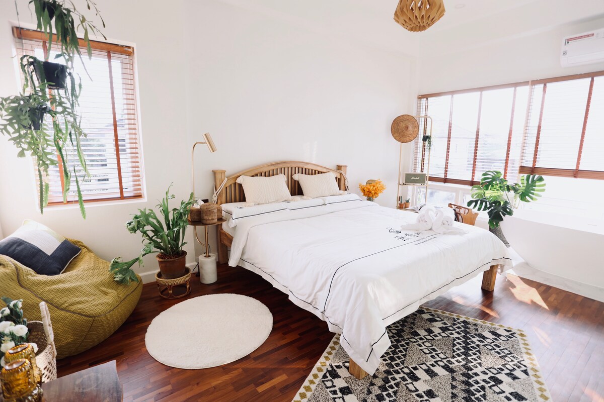A bright bedroom features a spacious bed with white bedding set against light walls. Natural light streams through large windows adorned with wooden blinds. A cozy bean bag chair, indoor plants, and a patterned rug add warmth to the inviting space.