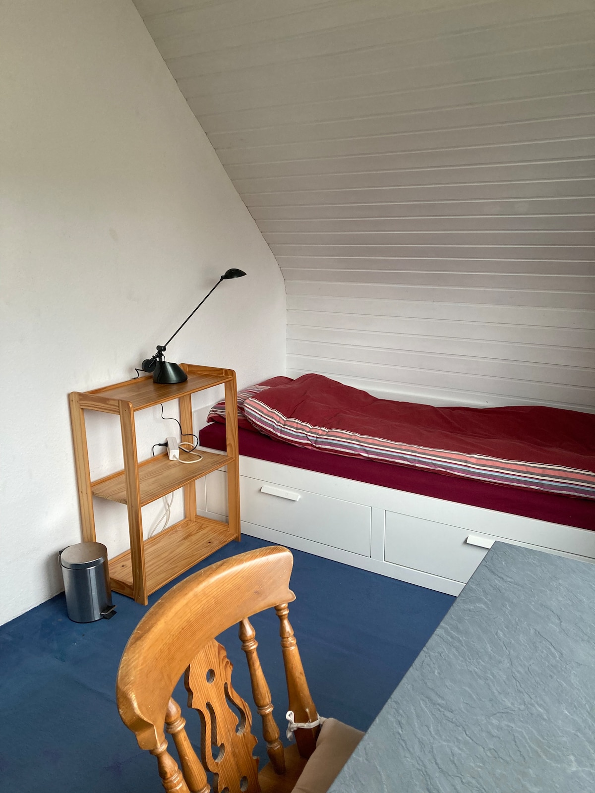 A cozy sleeping area features a narrow bed with a burgundy blanket. A wooden chair is placed at a small table, while a light fixture is mounted on a shelf beside the bed. The room is accented by slanted white walls and a blue carpet.