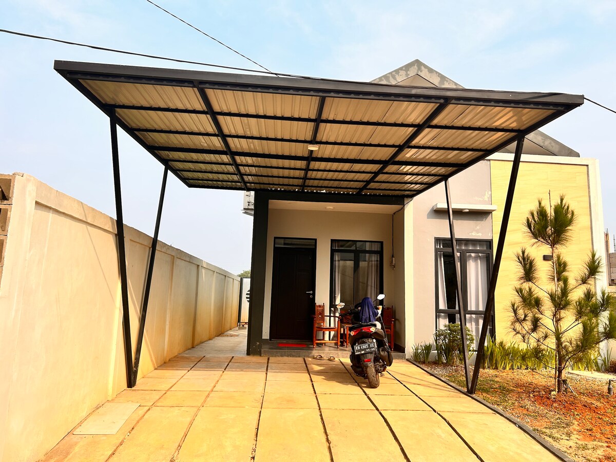 A covered parking area is presented in front of a modern residence, featuring a flat roof supported by sleek black beams. A motorcycle is parked beside a paved path leading to the entrance, which is flanked by low greenery and a privacy wall.