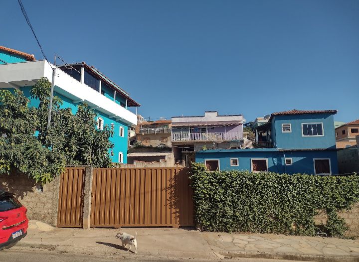 A multi-colored building exterior is visible, featuring a combination of blue and white walls. A wooden gate provides access to the property, framed by a green hedge. The surrounding area is characterized by a sunny sky and a peaceful neighborhood.