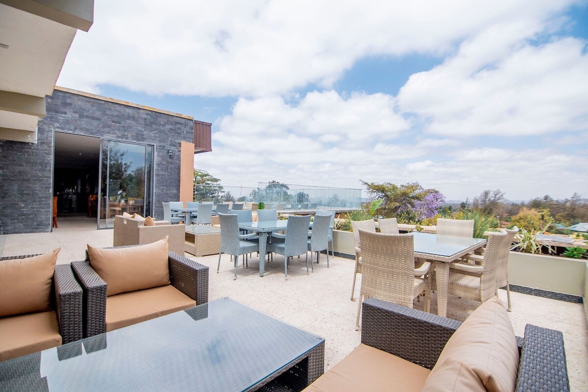 An outdoor seating area is highlighted by multiple tables and chairs arranged neatly on a patio. Comfortable lounge furniture is positioned alongside glass-top tables, providing a relaxed space for guests. The backdrop features trees and an expansive sky with scattered clouds.