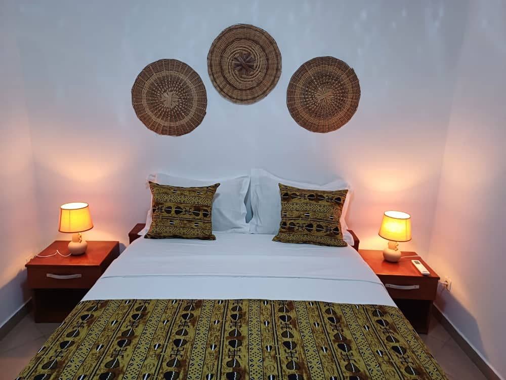 A cozy bedroom features a spacious bed adorned with patterned cushions and a decorative bedspread. Two bedside lamps cast a warm glow, complemented by woven wall art above the bed, enhancing the room's calming ambiance.