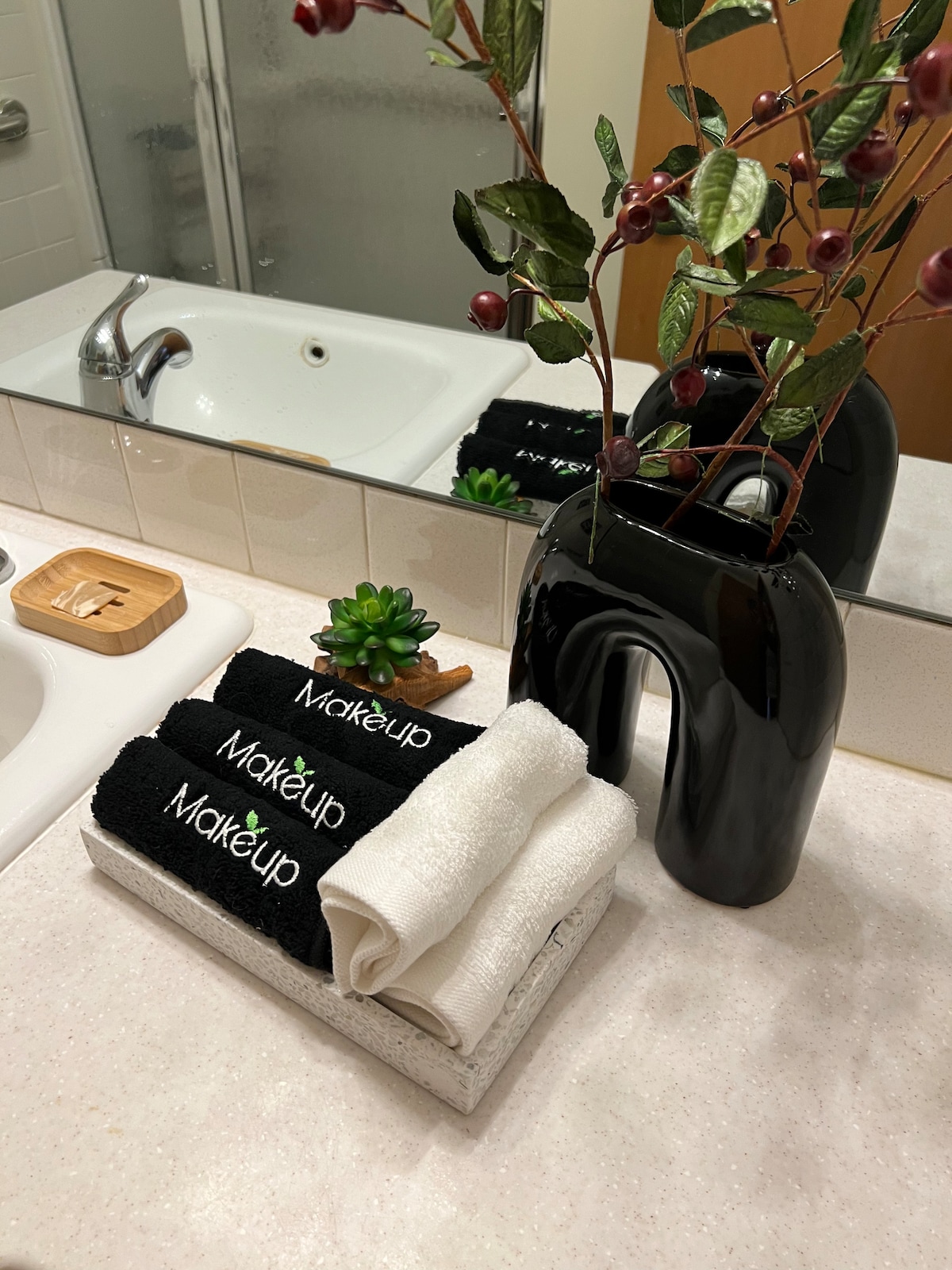 A bathroom countertop is displayed with neatly arranged towels in black and white, labeled with 'Makeup.' A small succulent plant and a decorative black vase filled with foliage add a touch of color. The reflection of the sink and mirror is visible in the background.