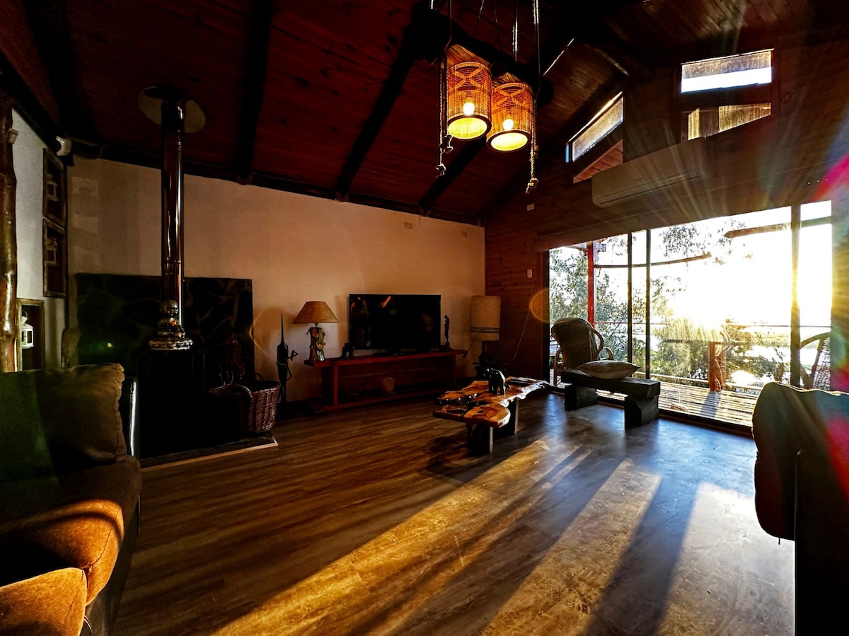 A spacious living area is highlighted by large windows that allow natural light to flow in, creating an airy ambiance. Cozy seating arrangements are present, featuring a plush sofa and lounge chairs. A flat-screen TV is positioned on a wooden stand, while a fireplace adds warmth to the space.