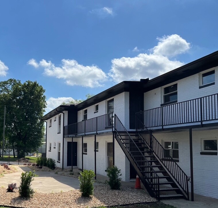 The exterior of a two-story building is shown, featuring a light-coloured facade and a staircase leading to the upper level. Nearby, landscaped areas with small shrubs are visible, and a clear blue sky with a few clouds completes the scene.