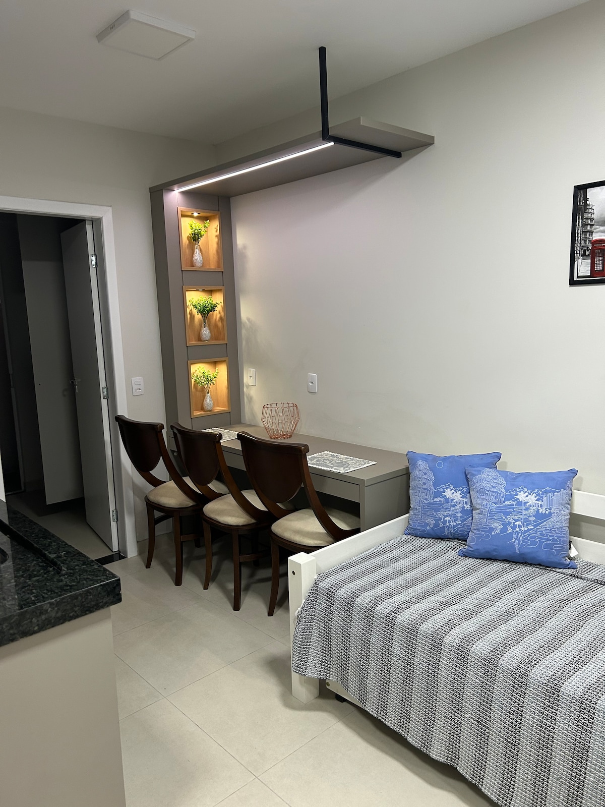 A cozy dining area is visible, featuring a table with four wooden chairs and decorative shelves illuminated with soft lighting. A comfortable seating space with a lightweight blanket lies against the wall, complemented by two decorative pillows. The overall layout suggests a welcoming and functional living space.