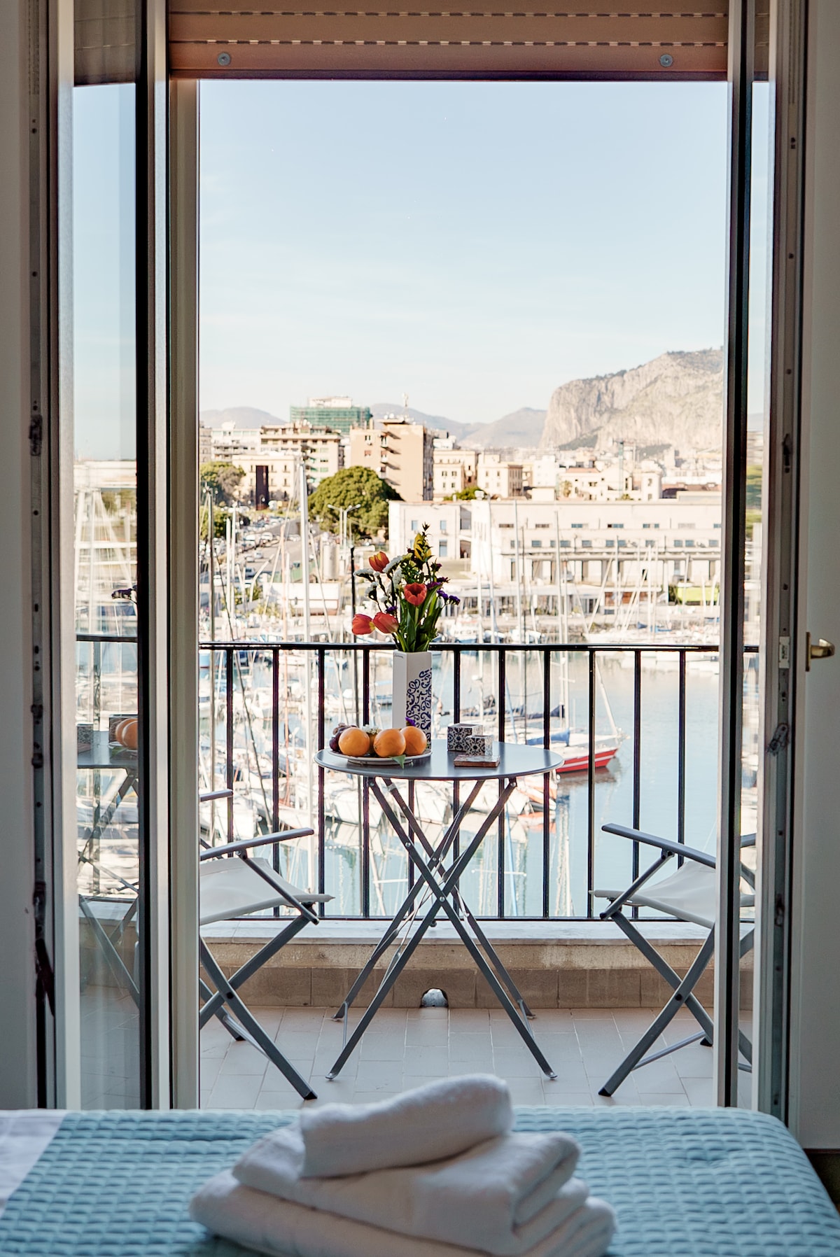 A balcony view is presented with a small round table and two chairs overlooking the marina. A floral arrangement and oranges are displayed on the table. The water reflects the surrounding buildings and hills, capturing the scenic beauty of the area.