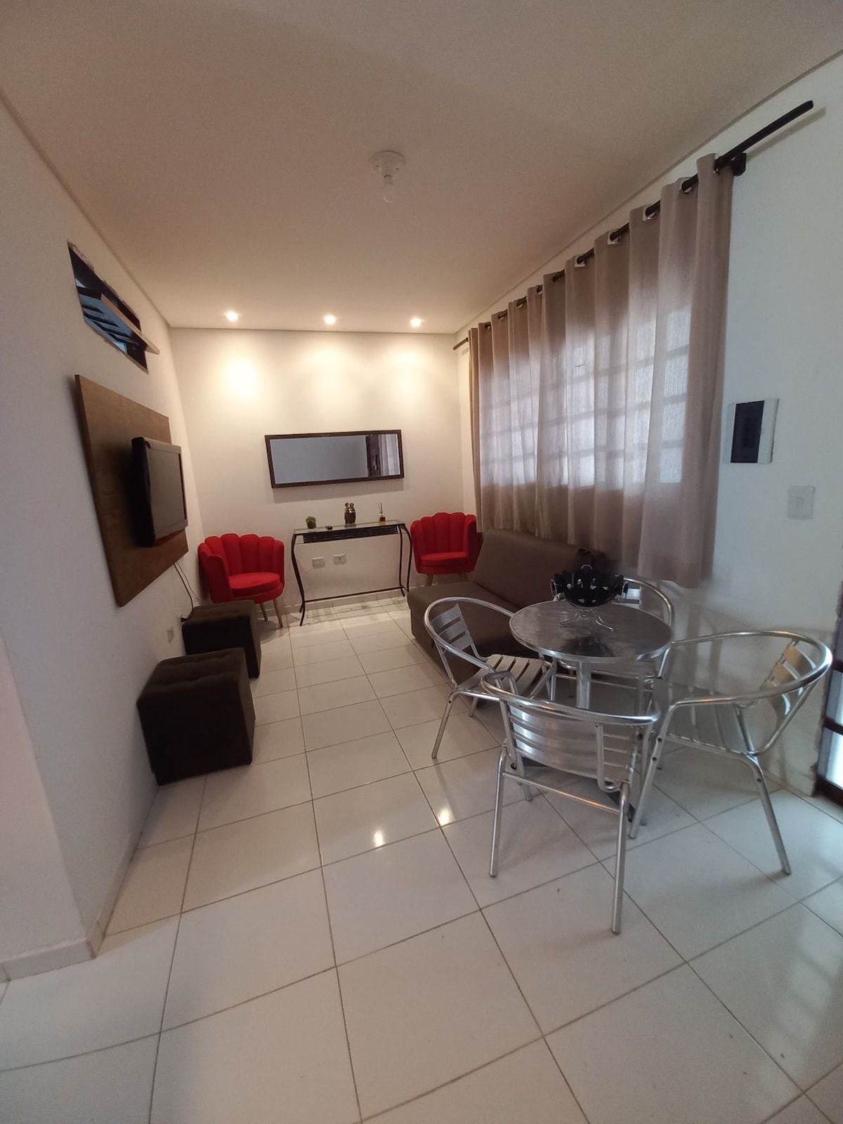 A modern living area features a sleek glass table surrounded by silver chairs, with two red accent chairs positioned nearby. A wall-mounted television is visible, and natural light filters through sheer curtains, creating a bright and open ambiance.