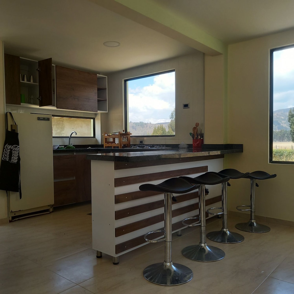 A modern kitchen features a sleek island with four bar stools. Large windows fill the space with natural light while offering views of the surrounding countryside. Cabinets and appliances are neatly arranged along one wall, creating an organized and functional cooking area.