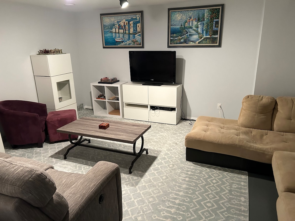 A cozy living room is displayed with a plush sectional sofa in neutral tones, complemented by two armchairs in rich burgundy. A wooden coffee table with a decorative item sits at the center. Entertainment options include a wall-mounted television and a white furniture unit, finished with framed artwork.