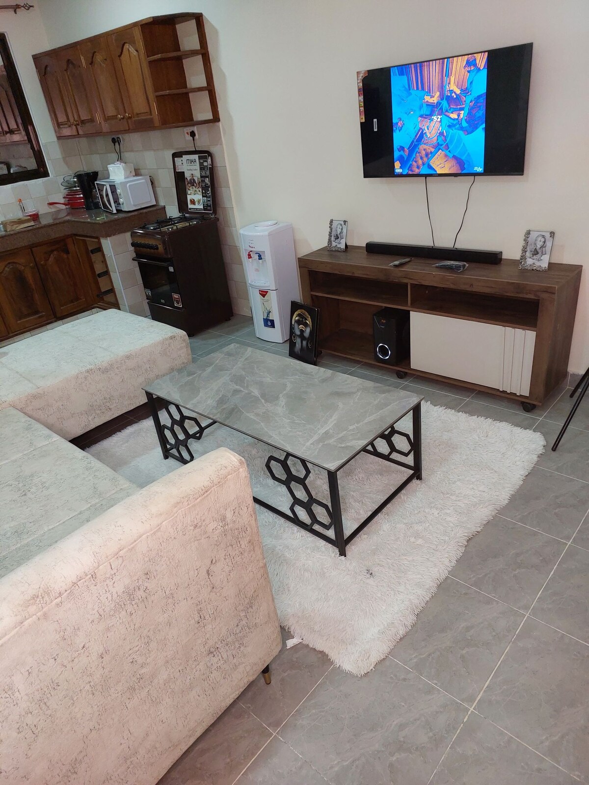A comfortable living area features two light-colored sofas facing a modern coffee table with a geometric design. A TV is mounted on the wall above a media console, and a rug adds softness to the tiled floor.