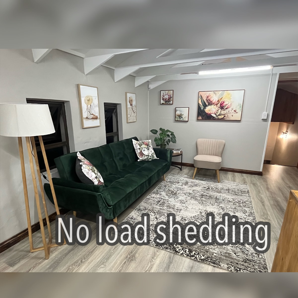 A spacious living area features a green velvet couch adorned with decorative pillows, complemented by a cozy armchair. Decorative wall art enhances the ambiance, while a patterned area rug anchors the seating arrangement. Natural light streams through small windows, illuminating the warm tones of the space.