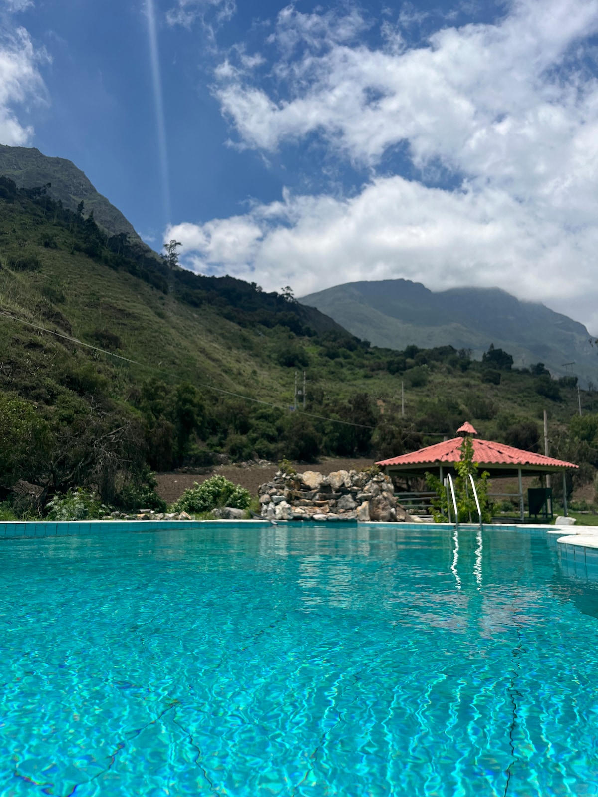 A clear blue pool reflects the surrounding mountains and sky. A sheltered seating area with a red tiled roof is visible beside the pool, framed by lush greenery, creating a serene outdoor space.