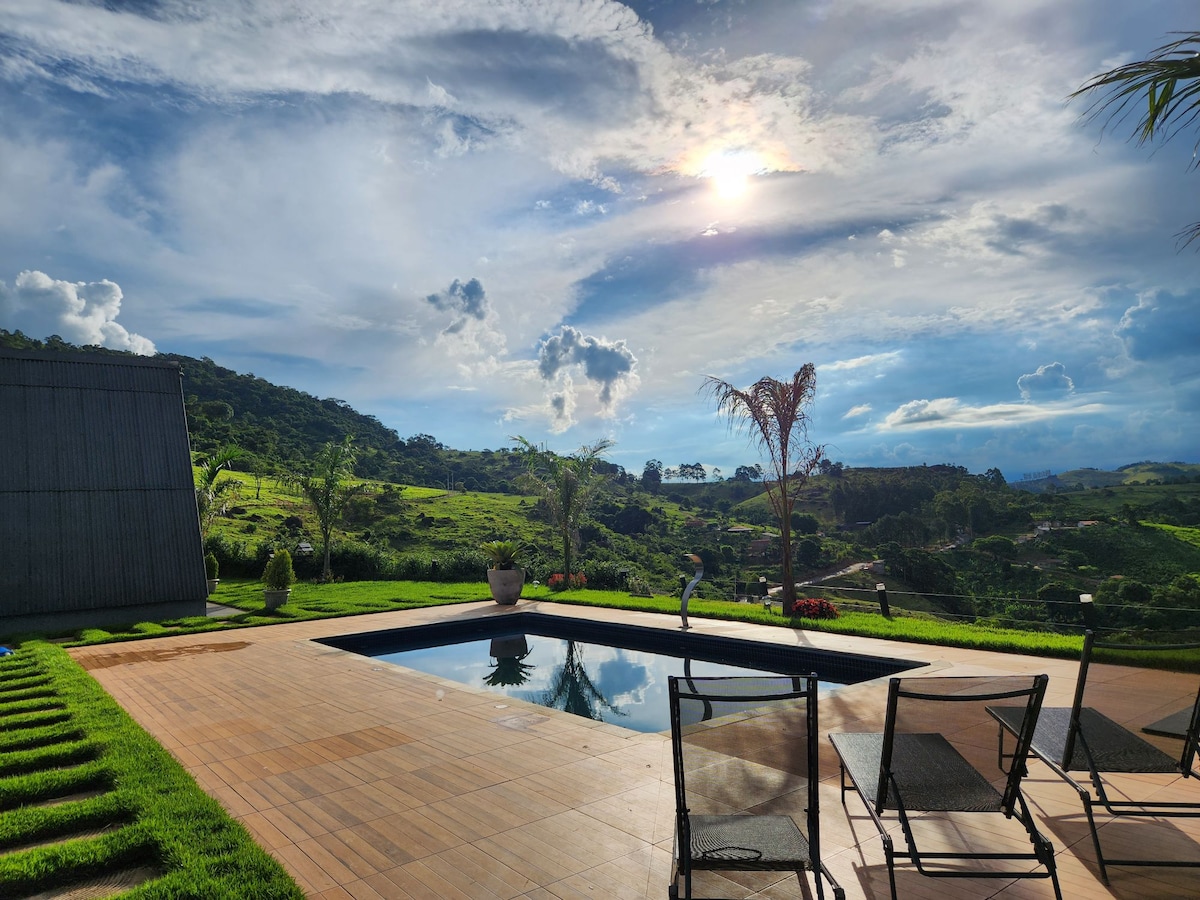A serene outdoor area features a sleek swimming pool surrounded by wooden decking. Lush green hills are visible in the background, under a sky with scattered clouds. Several black chairs are arranged around the pool, inviting relaxation.