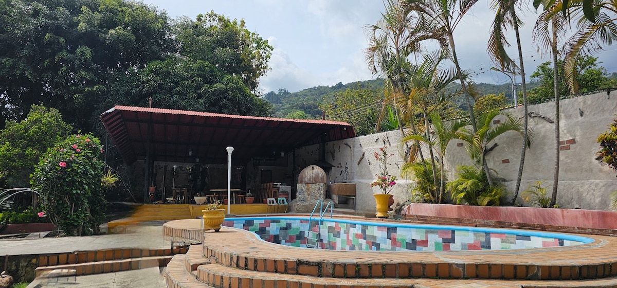 A spacious outdoor area features a swimming pool surrounded by vibrant greenery and palm trees. A covered seating area is visible with tables and chairs, providing shade. The backdrop showcases distant mountains, enhancing the natural setting.